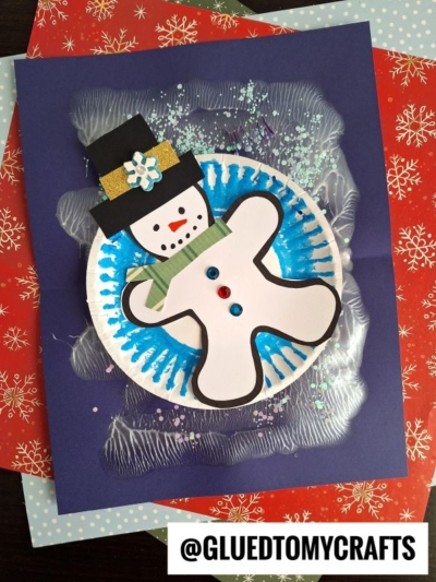 A handmade winter PAINT SPLAT CRAFT featuring a gingerbread man decorated as a snowman on a blue background, surrounded by blue and white paint, glitter, and layered festive papers. The @GLUEDTOMYCRAFTS tag is at the bottom.