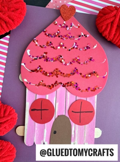 A Valentine-themed craft shaped like a house, made with pink popsicle sticks and a glittery red craft foam heart roof. Red circle windows, brown oval door, red pom-poms—perfect for heart shaped crafts or valentine art project ideas! @GLUEDTOMYCRAFTS at the bottom.