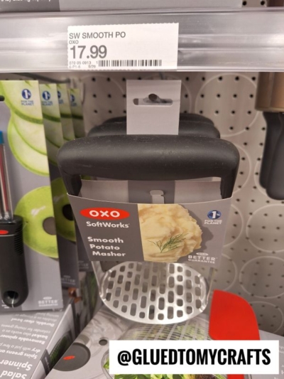 A stainless steel OXO SoftWorks smooth potato masher with a black handle is displayed on a store shelf. The price tag above shows $17.99. The Instagram handle @GLUEDTOMYCRAFTS is at the bottom.