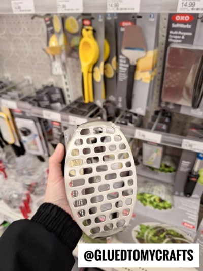 A hand holds a metal kitchen tool with a grid pattern over a store shelf displaying various OXO kitchen utensils and gadgets. The Instagram handle @GLUETOMYCRAFTS appears at the bottom.