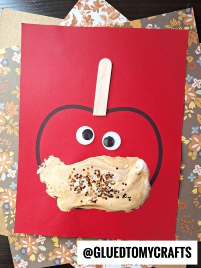 A red paper with a black outline of an apple, topped with a popsicle stick stem, two large googly eyes, and a peanut butter and sprinkles mix on the lower half, surrounded by fall-themed papers—perfect for pairing with puffy paint crafts.