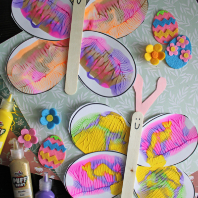 Two colorful easter egg butterfly crafts made from painted paper Easter eggs, with pink antennas and wooden craft stick bodies, are displayed on a floral background with felt flowers, painted eggs, and bottles of craft paint.