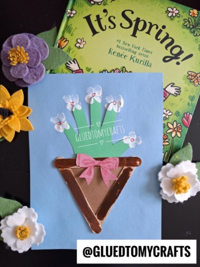 A spring-themed craft features a flower pot made from brown and gold paper, filled with white fingerprint flowers and a Popsicle Stick Flower Bouquet, topped with a pink bow, and displayed on blue paper. Felt flowers and a spring book decorate the background.