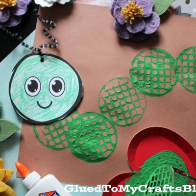 A close-up of a green caterpillar craft made with stamped green circles, a smiling paper face, felt flowers, and a bottle of glue. The blog name GluedToMyCraftsBlog.com appears at the bottom.