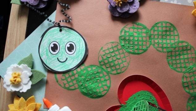 A close-up of a green caterpillar craft made with stamped green circles, a smiling paper face, felt flowers, and a bottle of glue. The blog name GluedToMyCraftsBlog.com appears at the bottom.
