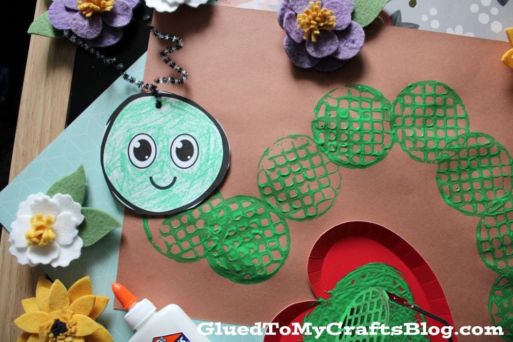 A close-up of a green caterpillar craft made with stamped green circles, a smiling paper face, felt flowers, and a bottle of glue. The blog name GluedToMyCraftsBlog.com appears at the bottom.