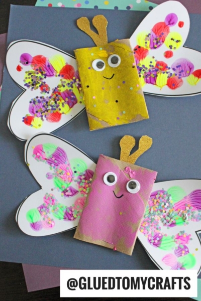 Two colorful butterfly INSECT CRAFT IDEAS made from decorated toilet paper rolls and paper wings, featuring googly eyes, smiley faces, painted patterns, and glittery antennas, displayed on a dark background.