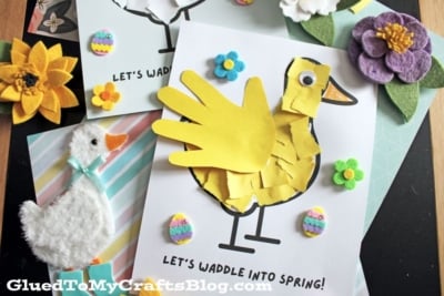 A spring-themed craft display features a yellow duck made from a child’s handprint, felt flowers, a felt duck, and colorful decorations on cards reading “Let’s waddle into spring!”.