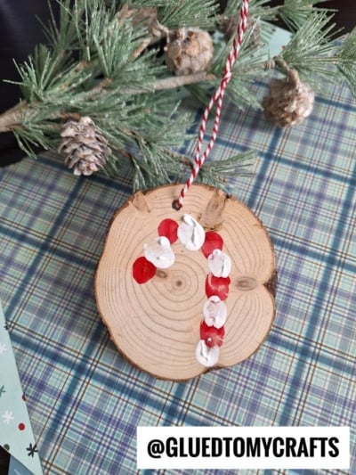A round wooden ornament with a red and white painted candy cane design hangs from a pine branch. Pinecones and plaid patterned paper are in the background. @GLUEDTOMYCRAFTS is written at the bottom.
