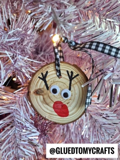 A wooden ornament painted as a reindeer face with googly eyes, red nose, and antler details hangs on a pink Christmas tree. A black-and-white ribbon is attached at the top. Text reads “@GLUEDTOMYCRAFTS.”.