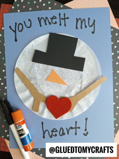 A handmade snowman card featuring coffee filter crafts for kids, with "You melt my heart!" written on blue paper. The snowman has brown paper arms, a red felt heart, and a black hat. Glue stick and markers are nearby. @GLUEDTOMYCRAFTS.
