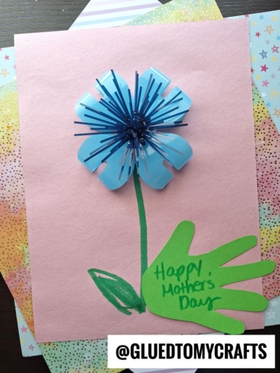 A handmade Mother’s Day card features a blue plastic flower with a green handprint stem and leaf, made from a childs cutout, on pink paper. The card says Happy Mother’s Day. @GLUEDTOMYCRAFTS is at the bottom.