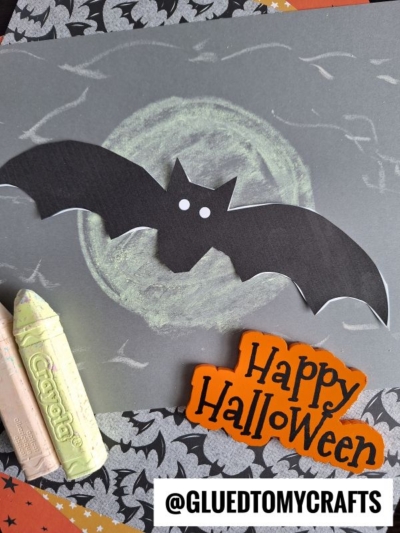 A Halloween craft featuring a black paper bat with white eyes on a chalk-drawn moon background, Crayola chalk, and a Happy Halloween sign—perfect for chalk art projects. Patterns of bats and stars border the image.