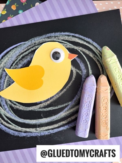 A yellow paper chick with a googly eye sits on black paper adorned with pastel chalk swirls, creating a fun chalk art project. Three Crayola chalks (yellow, purple, green) lie nearby, with @GLUEDTOMYCRAFTS at the bottom.
