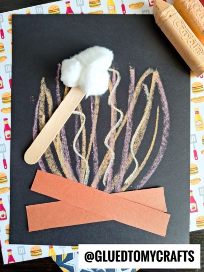 A craft of a campfire made with chalk flames on black paper, brown paper strips for logs, and a cotton ball on a craft stick for a marshmallow. Perfect for chalk art projects, with Crayola chalk nearby and a food-themed background pattern.
