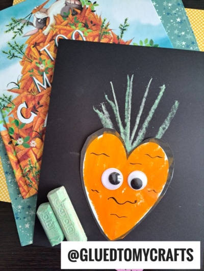A handmade carrot craft with googly eyes and a chalk-drawn green top is displayed on black paper, perfect for chalk art projects. Two sticks of green chalk and children's books about carrots sit nearby. The handle @GLUEDTOMYCRAFTS is shown at the bottom.