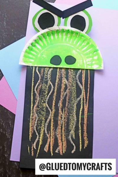 A frog craft made with a green painted paper plate, black paper, and pastel-colored squiggly lines on black paper for legs—perfect for chalk art projects—displayed on colored sheets. The handle @GLUEDTOMYCRAFTS is at the bottom.