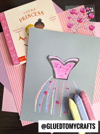 A princess dress craft made with pink and blue chalk on gray paper, decorated with sequins, surrounded by pink patterned papers, “A Little Princess” book, chalk pieces, a sheet of pink gems—perfect for chalk art projects.