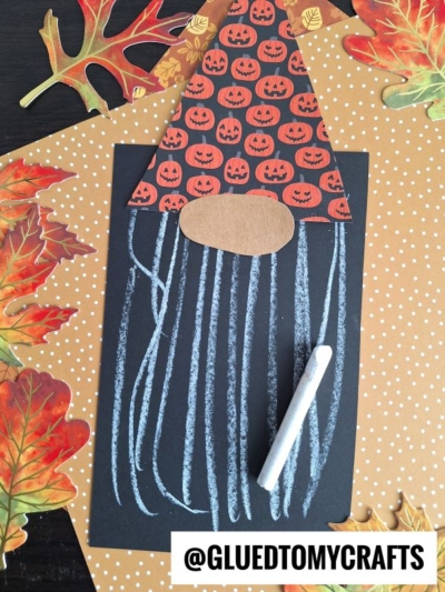 Halloween gnome craft made from black paper, a pumpkin-patterned hat, brown nose, and white chalk lines for a beard—perfect for chalk art projects. Surrounded by polka dot and autumn leaf papers, with a chalk piece and @GLUEDTOMYCRAFTS label.