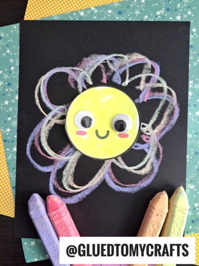 A children’s craft featuring a yellow smiling face with googly eyes on black paper, surrounded by colorful pastel chalk swirls—perfect for chalk art projects—with four large pastel crayons and a @GLUEDTOMYCRAFTS label at the bottom.