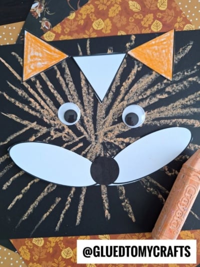 A fox face craft made with black paper, orange chalk art projects, googly eyes, and white and orange cut-out shapes, with the Instagram handle @gluedtomycrafts visible at the bottom. An orange crayon is nearby.