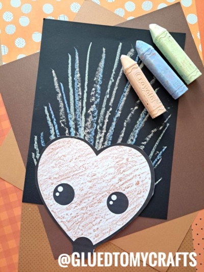 A heart-shaped paper hedgehog with large eyes sits on black paper, surrounded by brown and orange sheets. Spiky quills are drawn with pastel chalks—perfect for chalk art projects. Pastels sit nearby. Text reads @GLUEDTOMYCRAFTS.
