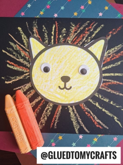 A yellow cat face with googly eyes and a drawn-on smile is centered on black paper with crayon starburst lines, perfect for chalk art projects. Two orange Crayola crayons and layered star-patterned papers are nearby. Text reads “@GLUEDTOMYCRAFTS.”.