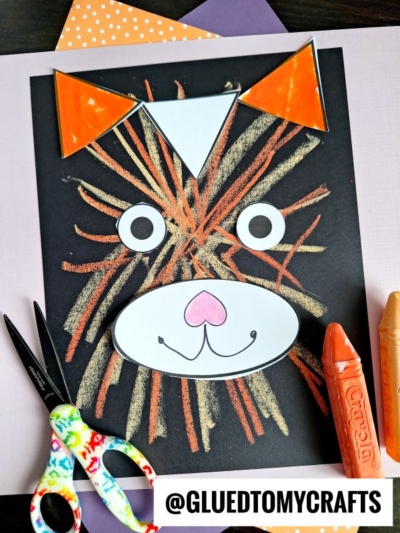 A cat face created on black paper using colored chalk for fur—a fun idea for chalk art projects—with orange paper ears, white eyes and muzzle, and a pink nose. Scissors, glue, and crayons are nearby. @GLUEDTOMYCRAFTS is at the bottom.