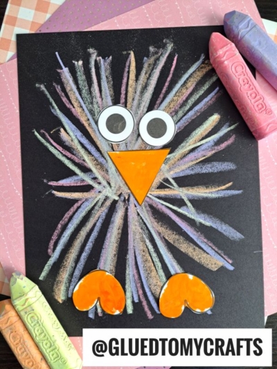A vibrant chalk art project, this colorful bird craft is made with pastel crayons on black paper, featuring googly eyes, an orange paper beak and feet. Large pastel crayons rest nearby. Instagram handle @GLUEDTOMYCRAFTS appears at the bottom.