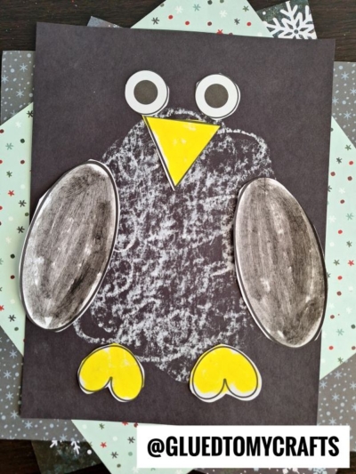 A handmade penguin craft using cut-out paper shapes for wings, beak, feet, and eyes is glued to a black background with a textured white oval for the body—perfect inspiration for chalk art projects. The username @GLUETOMYCRAFTS appears at the bottom.