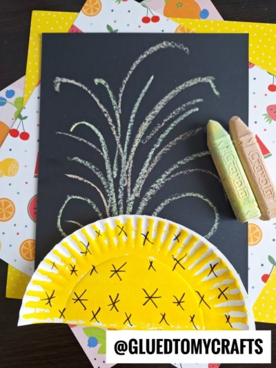 A black paper features chalk art projects with fireworks bursting from a yellow painted paper plate, resembling a pineapple half. Two pieces of chalk and colorful fruit-patterned paper are nearby. Text at the bottom reads @GLUEDTOMYCRAFTS.