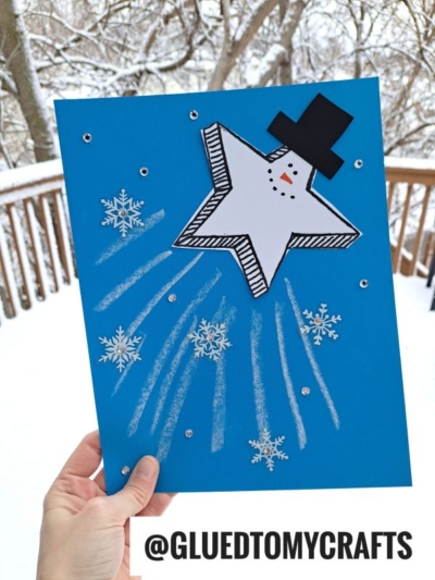 A hand holds a blue sheet of paper featuring a snowman star craft, chalk art project details like white chalk lines, snowflake stickers, and small silver dots. Snowy trees and a wooden fence appear in the background. @GLUEDTOMYCRAFTS is at the bottom.