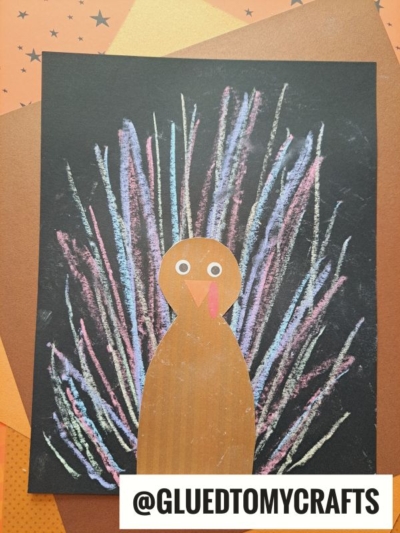 A turkey craft made with brown paper for the body and colorful chalk art project lines on black paper for feathers, placed on orange and brown star-patterned backgrounds. @GLUEDTOMYCRAFTS is written at the bottom.