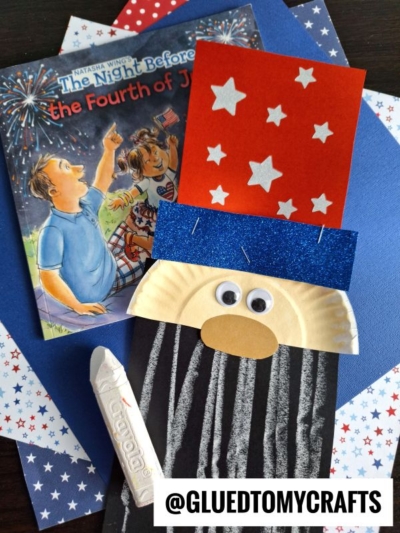 A children’s book about the Fourth of July sits beside a blue and red Abraham Lincoln craft made from a paper plate, googly eyes, and a popsicle stick, with star-patterned paper, chalk art projects, and a white crayon in the background.