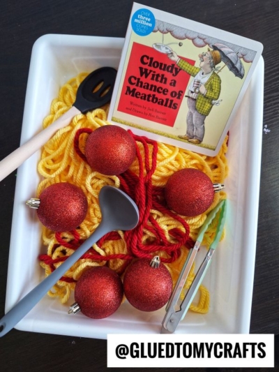 A white tray holds yellow yarn, red glittery balls, plastic utensils, tongs, and a Cloudy With a Chance of Meatballs book. @GLUEDTOMYCRAFTS is written at the bottom.