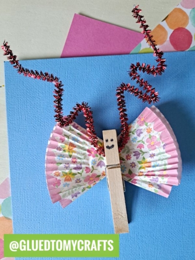 A butterfly craft from INSECT CRAFT IDEAS made with a clothespin, floral cupcake liners for wings, and red sparkly pipe cleaners for antennae sits on a blue paper background with colorful papers underneath.