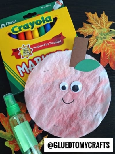 A pink coffee filter apple craft with googly eyes, a paper stem, and leaf sits next to a Crayola marker pack, spray bottle, and faux autumn leaves—perfect inspiration for coffee filter crafts for kids. Handle @GLUEDTOMYCRAFTS is at the bottom.