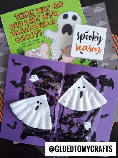 Children’s Halloween craft scene with a book titled “There Was an Old Lady Who Swallowed a Ghost!”, coffee filter crafts for kids like ghost crafts, purple paper bats, and a card reading “spooky season.”.