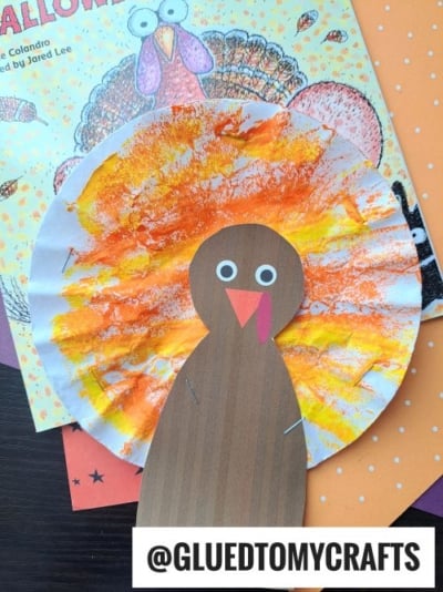 A paper turkey craft with a brown body and a colorful, painted coffee filter for feathers sits on a book and orange paper. Perfect for coffee filter crafts for kids! Feathers are yellow and orange. Text reads @GLUEDTOMYCRAFTS.