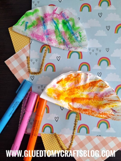 Two colorful paper plate rainbows with gold pipe cleaner handles are displayed on pastel rainbow-themed paper, alongside three markers. Perfect for coffee filter crafts for kids to spark creativity and brighten any day.