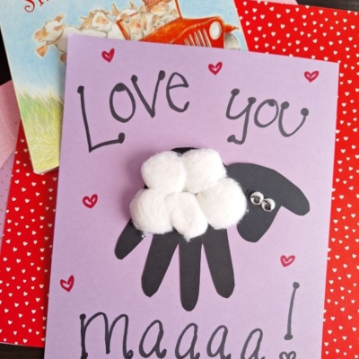 A handmade card reads Love you maaaa! with a black handprint sheep made of cotton balls and googly eyes. The card sits on red and pink paper and a childrens book titled Sheep in a Jeep.