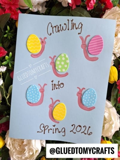 A blue card decorated with colorful egg-shaped snails and the handwritten text Crawling into Spring 2026. The card is surrounded by flowers, and @GLUEDTOMYCRAFTS is displayed at the bottom.
