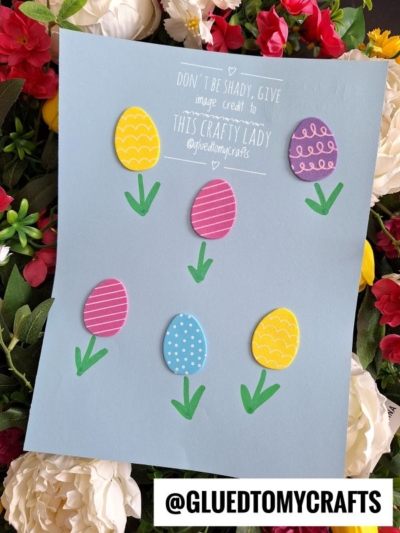 A blue sheet of paper displays six colorful paper Easter eggs with drawn patterns and green stems, arranged among faux flowers. Text at the top credits the crafter, and @GLUEDTOMYCRAFTS is labeled at the bottom.