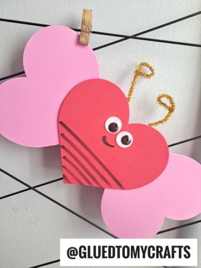 A heart-shaped butterfly made from craft foam heart crafts in pink and red, with googly eyes, a smile, gold pipe cleaner antennae, and pink wings—perfect for those seeking unique valentine art project ideas. Text at the bottom reads @GLUEDTOMYCRAFTS.