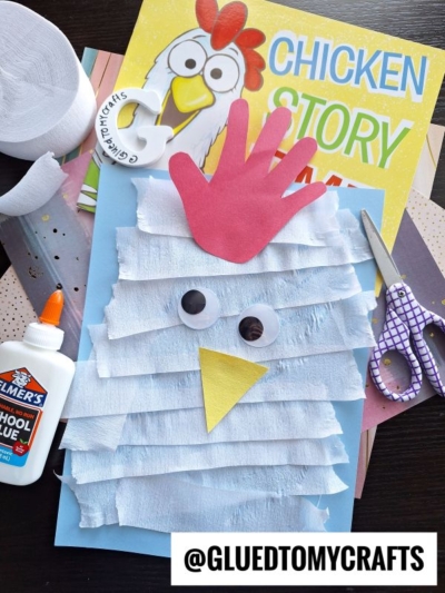 A fun crepe paper craft idea: a children’s chicken made with white crepe paper strips, a red handprint comb, googly eyes, and a yellow beak sits on blue paper. Craft supplies and a “Chicken Story” book are nearby.