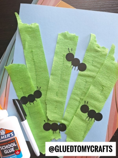 A creative project features green crepe paper craft ideas, with streamer strips on blue paper and black circles forming ants. A glue bottle and black marker rest nearby. The text @GLUEDTOMYCRAFTS appears at the bottom.