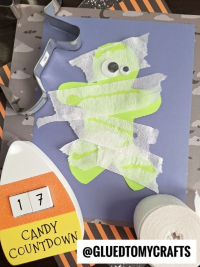 A green paper cutout shaped like a person is decorated with white crepe paper strips and a googly eye to resemble a mummy—a fun addition to your crepe paper craft ideas. Nearby are a candy countdown sign, craft supplies, and a roll of crepe paper.