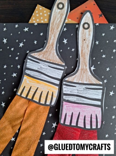 Two paper paintbrush crafts with brown handles and colored bristles, decorated with crepe paper craft ideas using orange and red tissue paper, are placed on a star-patterned background. The text @GLUEDTOMYCRAFTS appears at the bottom.