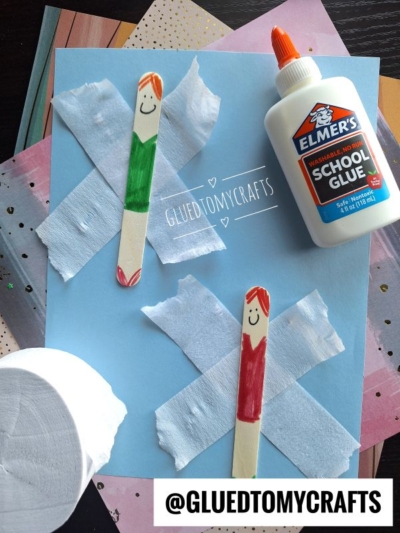 Two popsicle stick people, dressed in green and red, are glued to blue paper with white tissue and school glue—a fun example of crepe paper craft ideas. Craft materials and paper surround the artwork. Text reads @GLUEDTOMYCRAFTS.