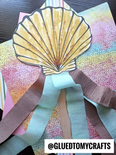A paper seashell craft with brown and blue crepe paper streamers hangs from the bottom, displayed on colorful patterned paper—perfect inspiration for your next crepe paper craft ideas. The handle @GLUEDTOMYCRAFTS is visible at the bottom.
